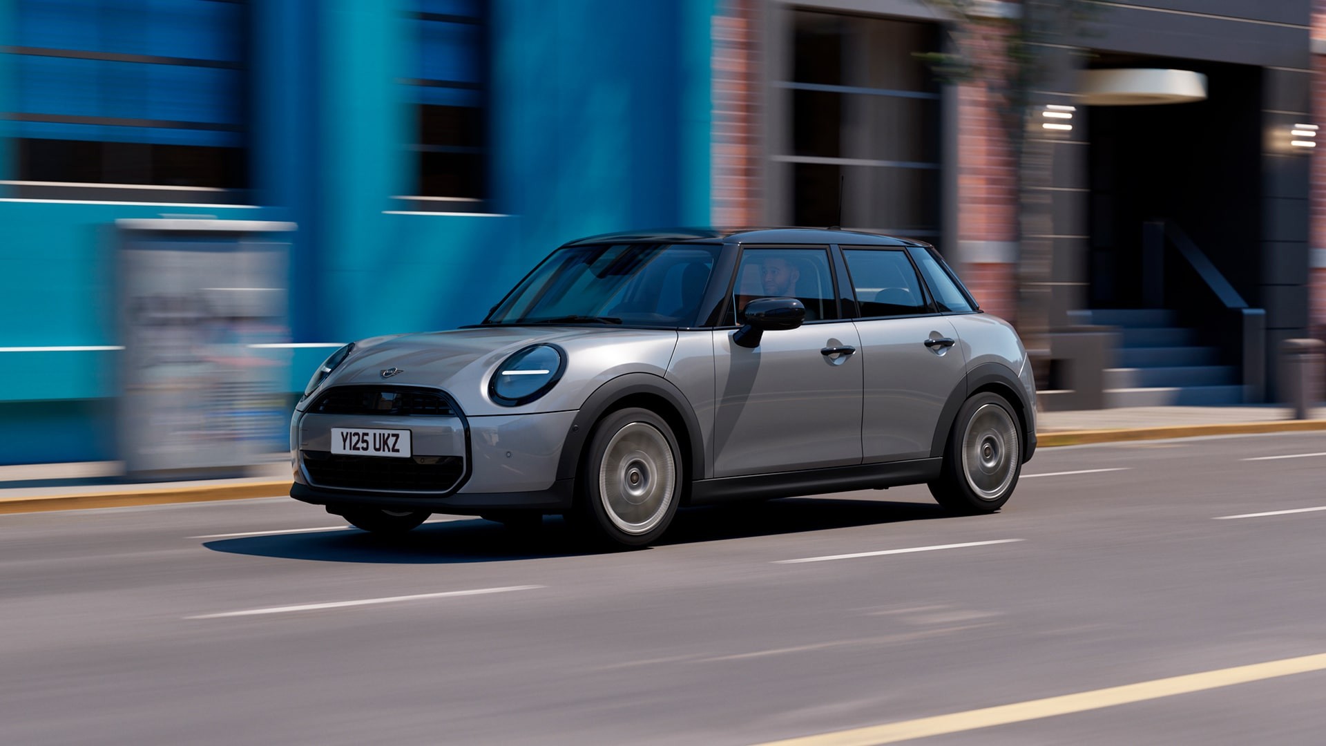 A silver MINI Cooper 5-Door drives swiftly down a city street, passing a bright blue wall and brick building. The scene conveys a sense of speed and urban energy.